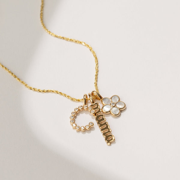 A textured gold chain with an initial 'C' charm made up of small round clear gemstones, a vertical gold charm with the word 'mama' in script font, and a daisy charm with a white stone making up the petals, laying on a solid white background with shadows.