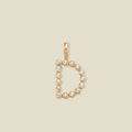 A gold letter 'D' pendant charm embellished with small round-cut clear gemstones, centered on a solid light beige background.