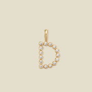 A gold letter 'D' pendant charm embellished with small round-cut clear gemstones, centered on a solid light beige background.