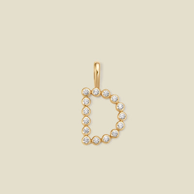 A gold letter 'D' pendant charm embellished with small round-cut clear gemstones, centered on a solid light beige background.