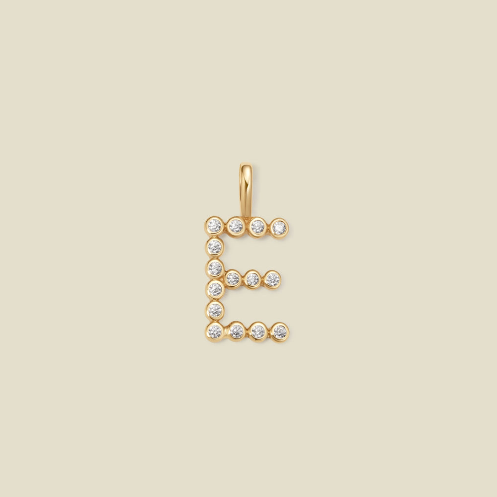 A gold letter 'E' pendant charm embellished with small clear round gemstones, centered on a solid light beige background.