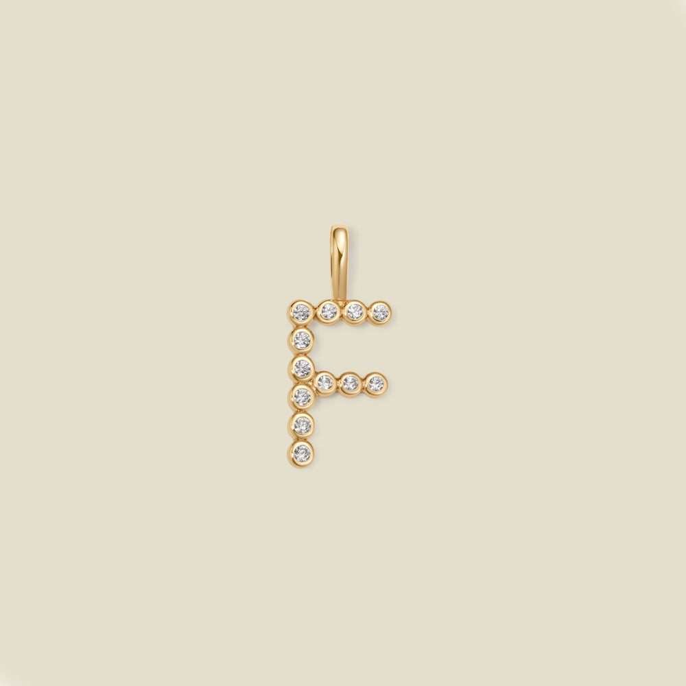 A gold letter 'F' pendant charm embellished with small clear round gemstones, centered on a solid light beige background.