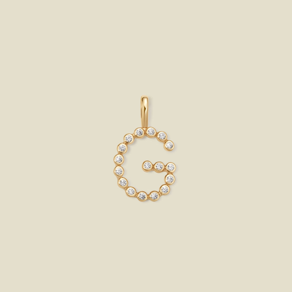 A gold letter 'G' pendant charm embellished with small clear round gemstones, centered on a solid light beige background.