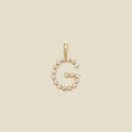 A gold letter 'G' pendant charm embellished with small clear round gemstones, centered on a solid light beige background.