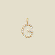 A gold letter 'G' pendant charm embellished with small clear round gemstones, centered on a solid light beige background.