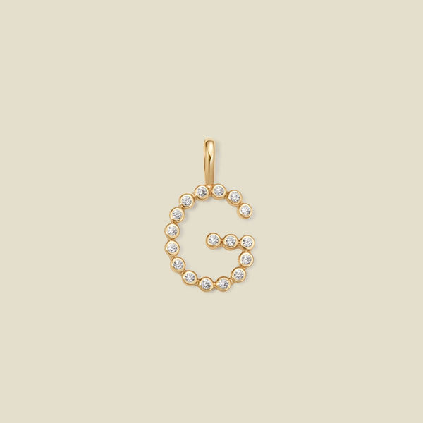 A gold letter 'G' pendant charm embellished with small clear round gemstones, centered on a solid light beige background.