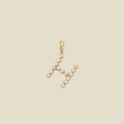 A gold letter 'H' pendant charm embellished with small clear gemstones, set against a solid light beige background.