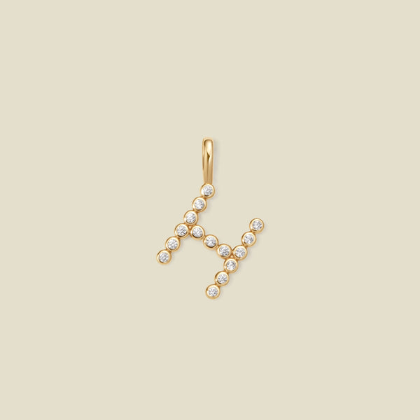 A gold letter 'H' pendant charm embellished with small clear gemstones, set against a solid light beige background.