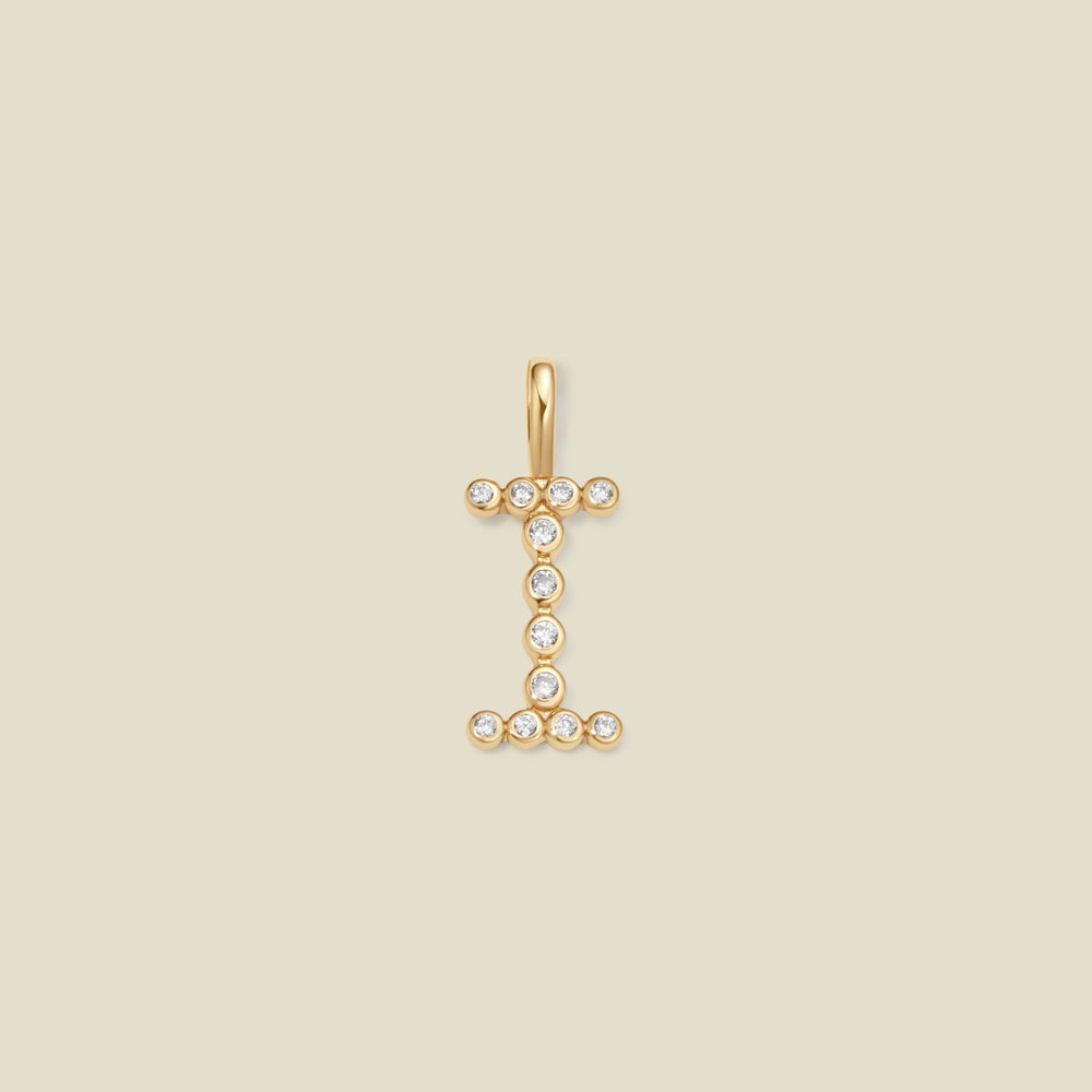 A gold initial pendant charm shaped like the letter 'I', adorned with small clear gemstones, centered on a solid beige background.