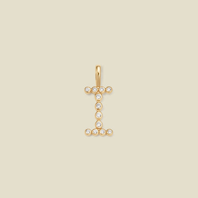 A gold initial pendant charm shaped like the letter 'I', adorned with small clear gemstones, centered on a solid beige background.