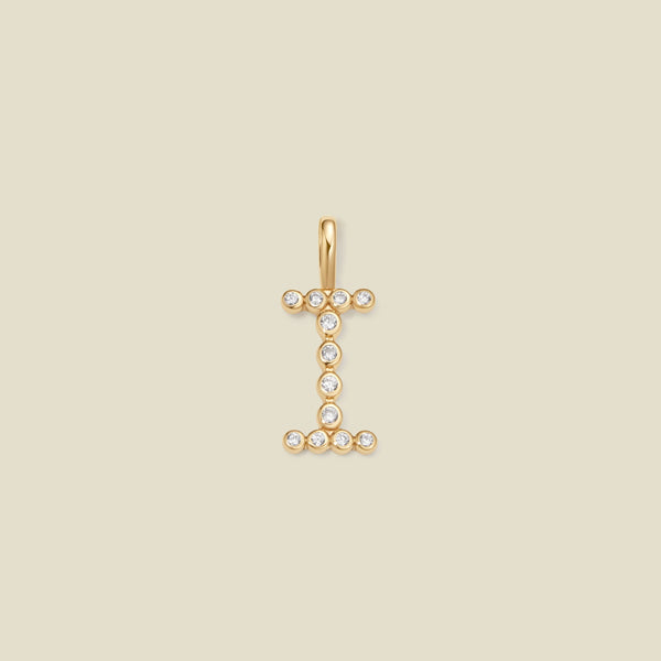 A gold initial pendant charm shaped like the letter 'I', adorned with small clear gemstones, centered on a solid beige background.