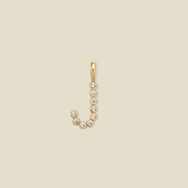 A gold letter 'J' initial pendant charm made up of clear round-cut bezel set gemstones, set against a solid light beige background.
