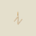 A gold initial 'N' charm with clear round gemstones, centered on a solid beige background.