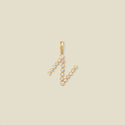 A gold initial 'N' charm with clear round gemstones, centered on a solid beige background.