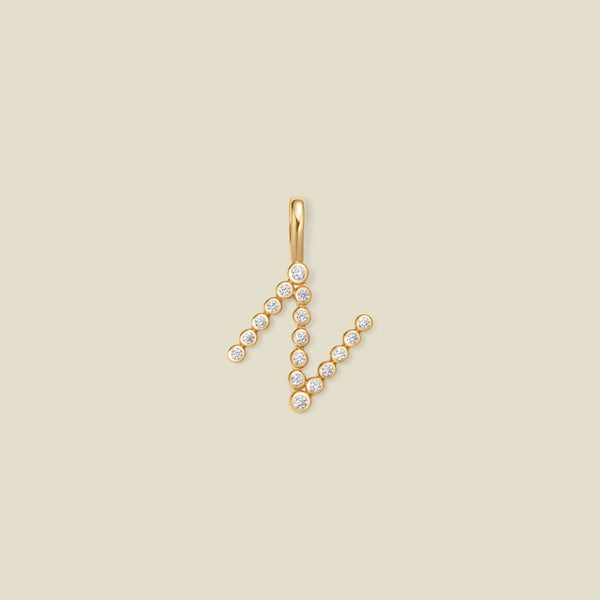 A gold initial 'N' charm with clear round gemstones, centered on a solid beige background.