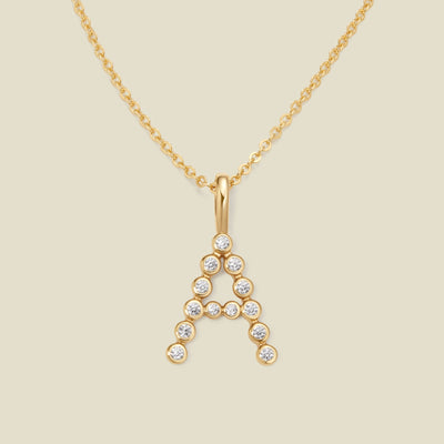 A gold pendant chain necklace with a gold uppercase letter 'A' charm made up of round bezel-set clear gemstones, set against a solid beige background.