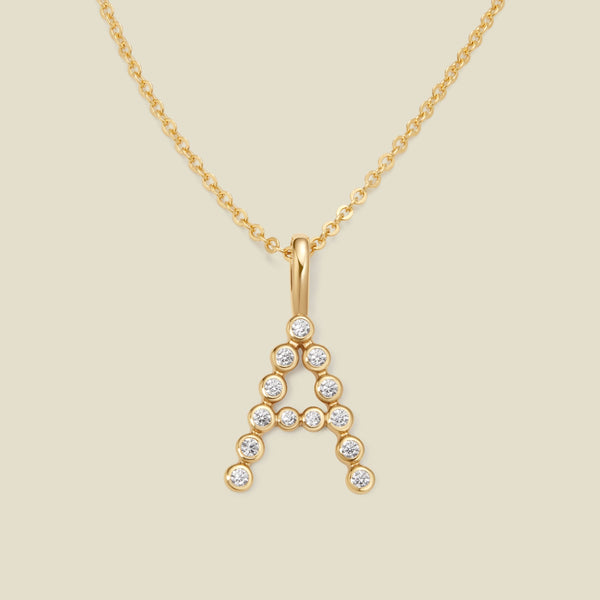 A gold pendant chain necklace with a gold uppercase letter 'A' charm made up of round bezel-set clear gemstones, set against a solid beige background.
