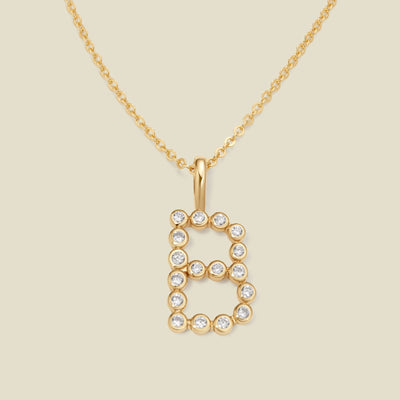 A gold pendant chain necklace with a gold uppercase letter 'B' charm made up of round bezel-set clear gemstones, set against a solid beige background.