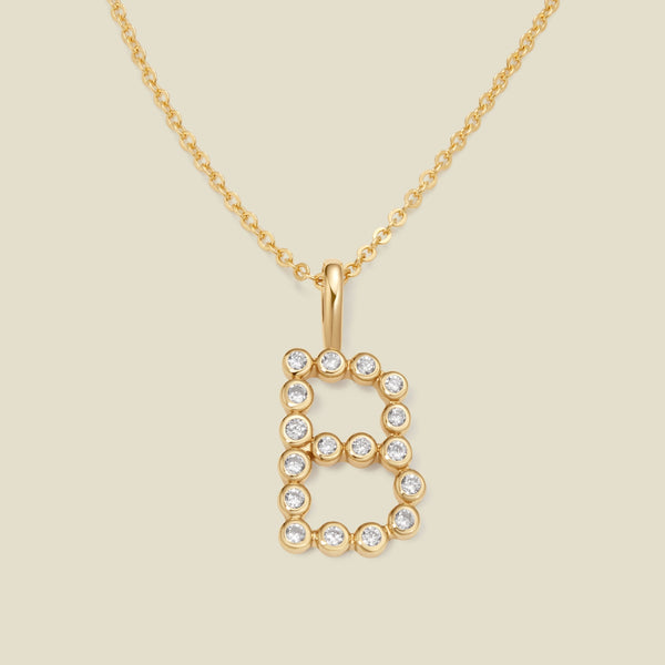 A gold pendant chain necklace with a gold uppercase letter 'B' charm made up of round bezel-set clear gemstones, set against a solid beige background.