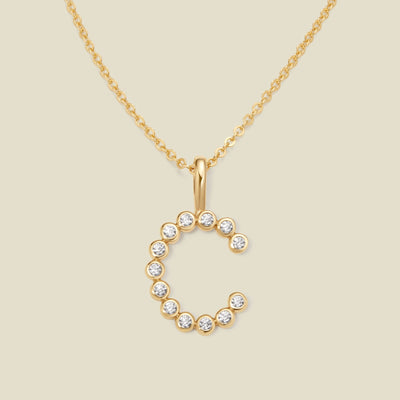 A gold pendant chain necklace with a gold uppercase letter 'C' charm made up of round bezel-set clear gemstones, set against a solid beige background.
