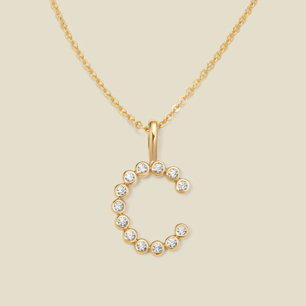 A gold pendant chain necklace with a gold uppercase letter 'C' charm made up of round bezel-set clear gemstones, set against a solid beige background.