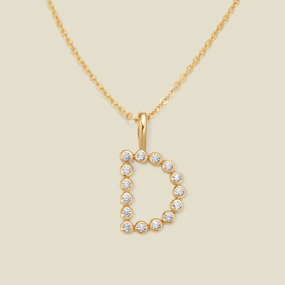 A gold pendant chain necklace with a gold uppercase letter 'D' charm made up of round bezel-set clear gemstones, set against a solid beige background.