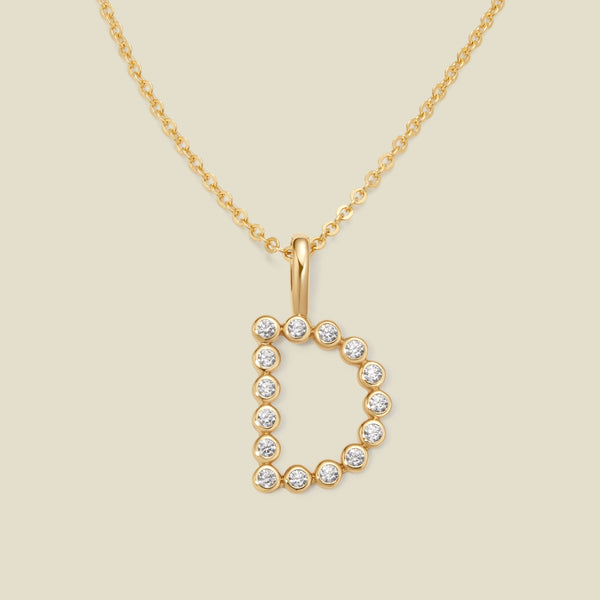 A gold pendant chain necklace with a gold uppercase letter 'D' charm made up of round bezel-set clear gemstones, set against a solid beige background.