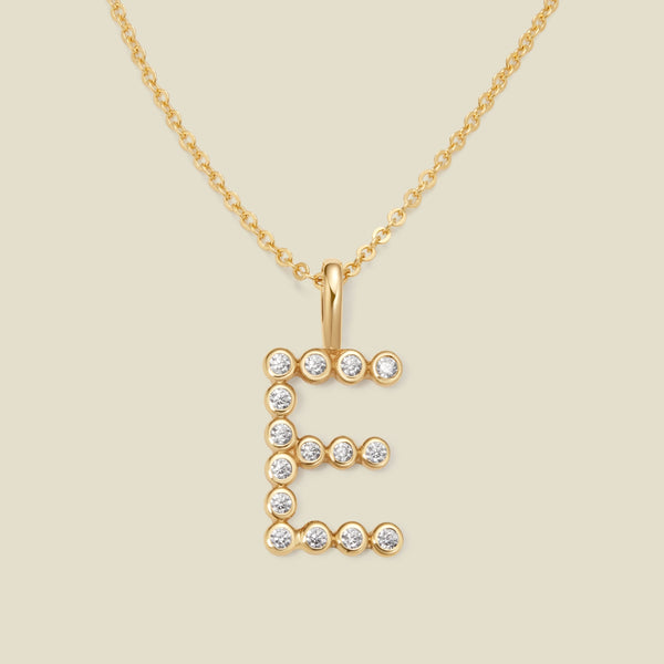 A gold pendant chain necklace with a gold uppercase letter 'E' charm made up of round bezel-set clear gemstones, set against a solid beige background.