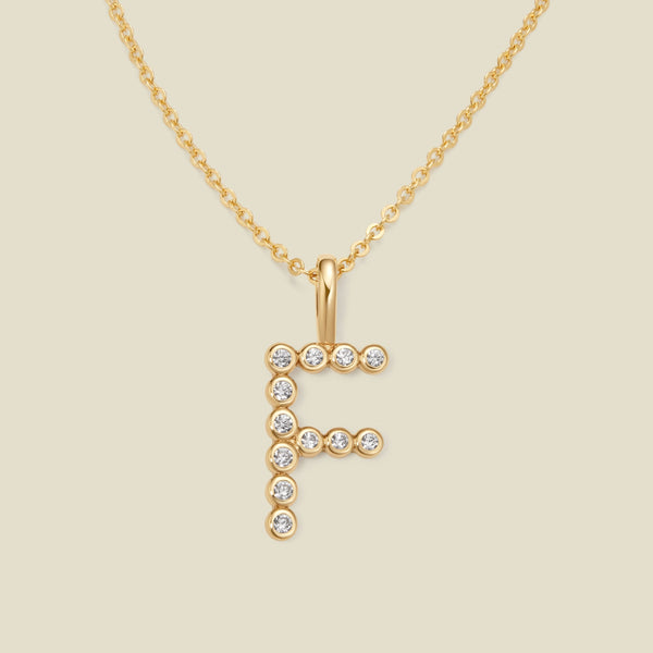 A gold pendant chain necklace with a gold uppercase letter 'F' charm made up of round bezel-set clear gemstones, set against a solid beige background.