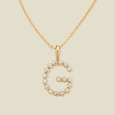 A gold pendant chain necklace with a gold uppercase letter 'G' charm made up of round bezel-set clear gemstones, set against a solid beige background.