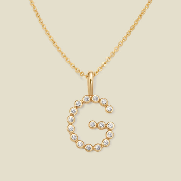 A gold pendant chain necklace with a gold uppercase letter 'G' charm made up of round bezel-set clear gemstones, set against a solid beige background.