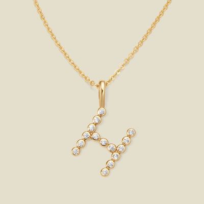 A gold pendant chain necklace with a gold uppercase letter 'H' charm made up of round bezel-set clear gemstones, set against a solid beige background.