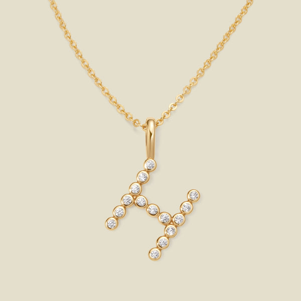 A gold pendant chain necklace with a gold uppercase letter 'H' charm made up of round bezel-set clear gemstones, set against a solid beige background.