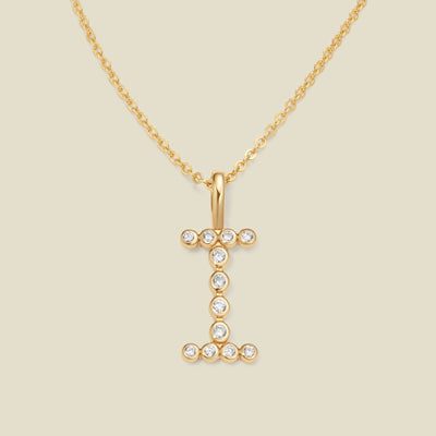 A gold pendant chain necklace with a gold uppercase letter 'I' charm made up of round bezel-set clear gemstones, set against a solid beige background.