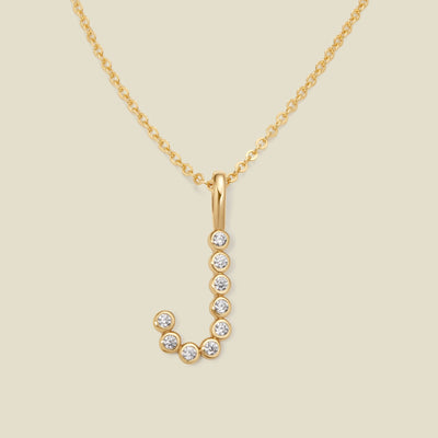 A gold pendant chain necklace with a gold uppercase letter 'J' charm made up of round bezel-set clear gemstones, set against a solid beige background.