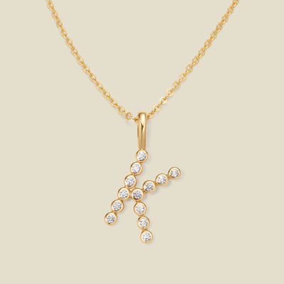 A gold pendant chain necklace with a gold uppercase letter 'K' charm made up of round bezel-set clear gemstones, set against a solid beige background.