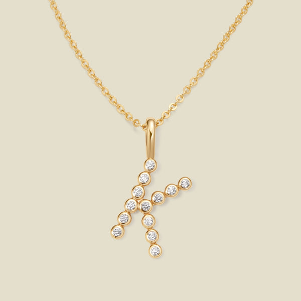 A gold pendant chain necklace with a gold uppercase letter 'K' charm made up of round bezel-set clear gemstones, set against a solid beige background.