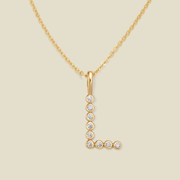 A gold pendant chain necklace with a gold uppercase letter 'L' charm made up of round bezel-set clear gemstones, set against a solid beige background.