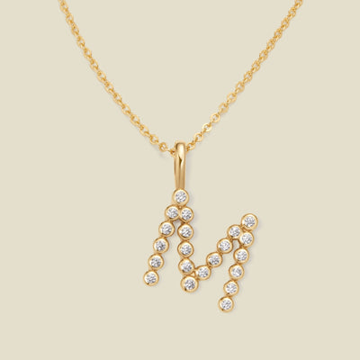 A gold pendant chain necklace with a gold uppercase letter 'M' charm made up of round bezel-set clear gemstones, set against a solid beige background.