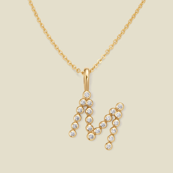 A gold pendant chain necklace with a gold uppercase letter 'M' charm made up of round bezel-set clear gemstones, set against a solid beige background.