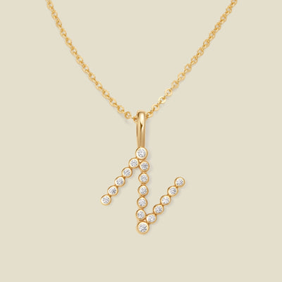 A gold pendant chain necklace with a gold uppercase letter 'N' charm made up of round bezel-set clear gemstones, set against a solid beige background.