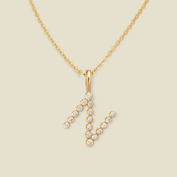 A gold pendant chain necklace with a gold uppercase letter 'N' charm made up of round bezel-set clear gemstones, set against a solid beige background.