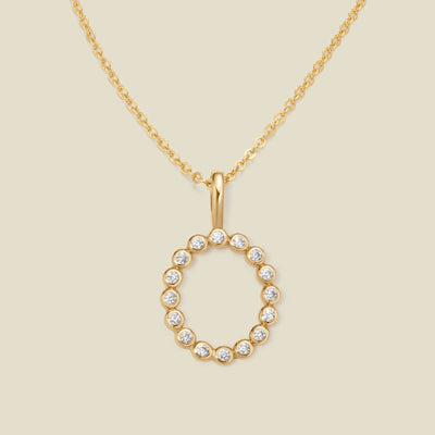 A gold pendant chain necklace with a gold uppercase letter 'O' charm made up of round bezel-set clear gemstones, set against a solid beige background.