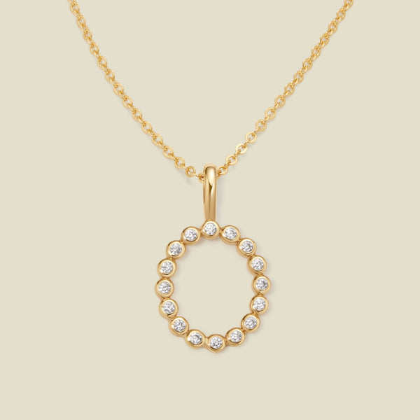A gold pendant chain necklace with a gold uppercase letter 'O' charm made up of round bezel-set clear gemstones, set against a solid beige background.