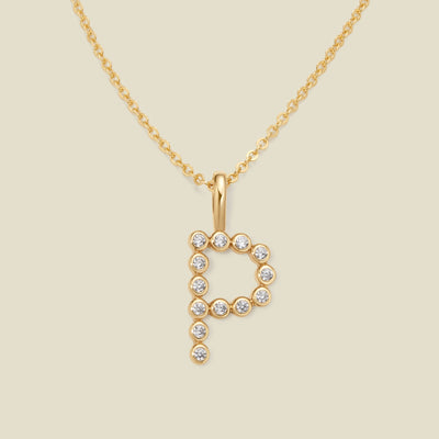 A gold pendant chain necklace with a gold uppercase letter 'P' charm made up of round bezel-set clear gemstones, set against a solid beige background.