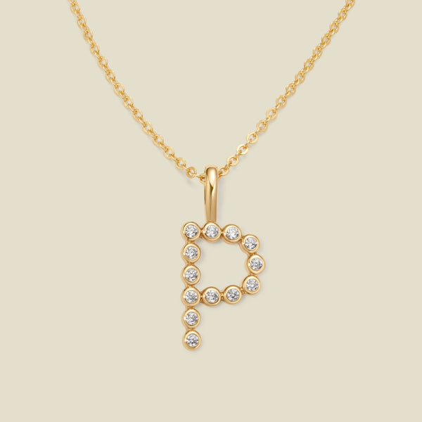 A gold pendant chain necklace with a gold uppercase letter 'P' charm made up of round bezel-set clear gemstones, set against a solid beige background.