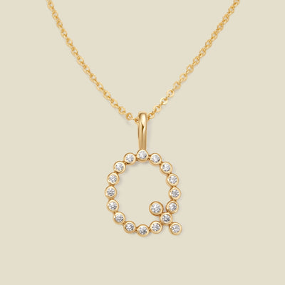 A gold pendant chain necklace with a gold uppercase letter 'Q' charm made up of round bezel-set clear gemstones, set against a solid beige background.