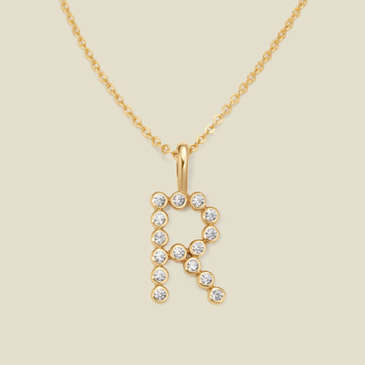 A gold pendant chain necklace with a gold uppercase letter 'R' charm made up of round bezel-set clear gemstones, set against a solid beige background.