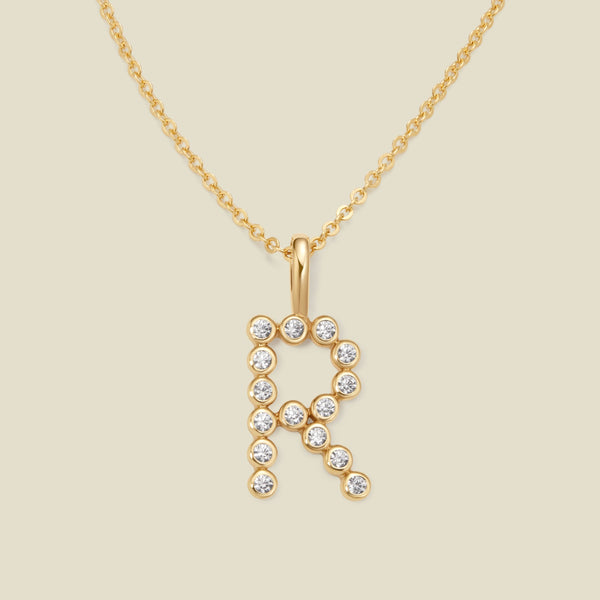 A gold pendant chain necklace with a gold uppercase letter 'R' charm made up of round bezel-set clear gemstones, set against a solid beige background.