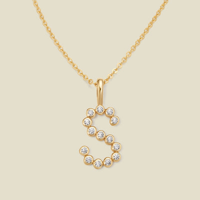 A gold pendant chain necklace with a gold uppercase letter 'S' charm made up of round bezel-set clear gemstones, set against a solid beige background.